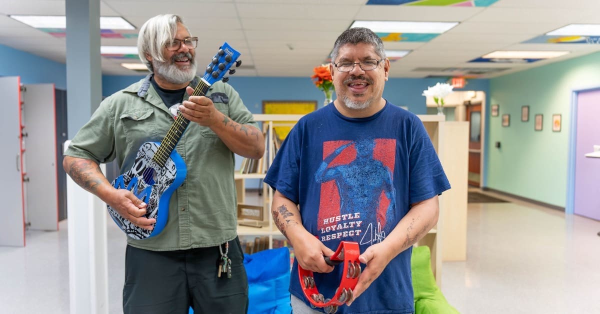 A TLC employee plays a guitar next to a TLC individual playing a tambourine during an indoor music party.