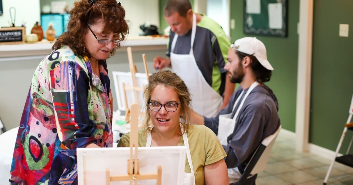 An art teacher stands next to a TLC individual behind a paint canvas.