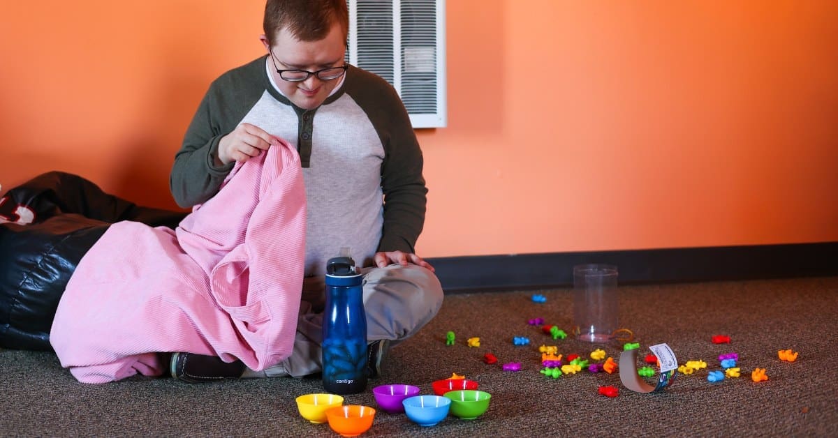 A TLC individual sits on the floor while playing a game with colorful bowls.