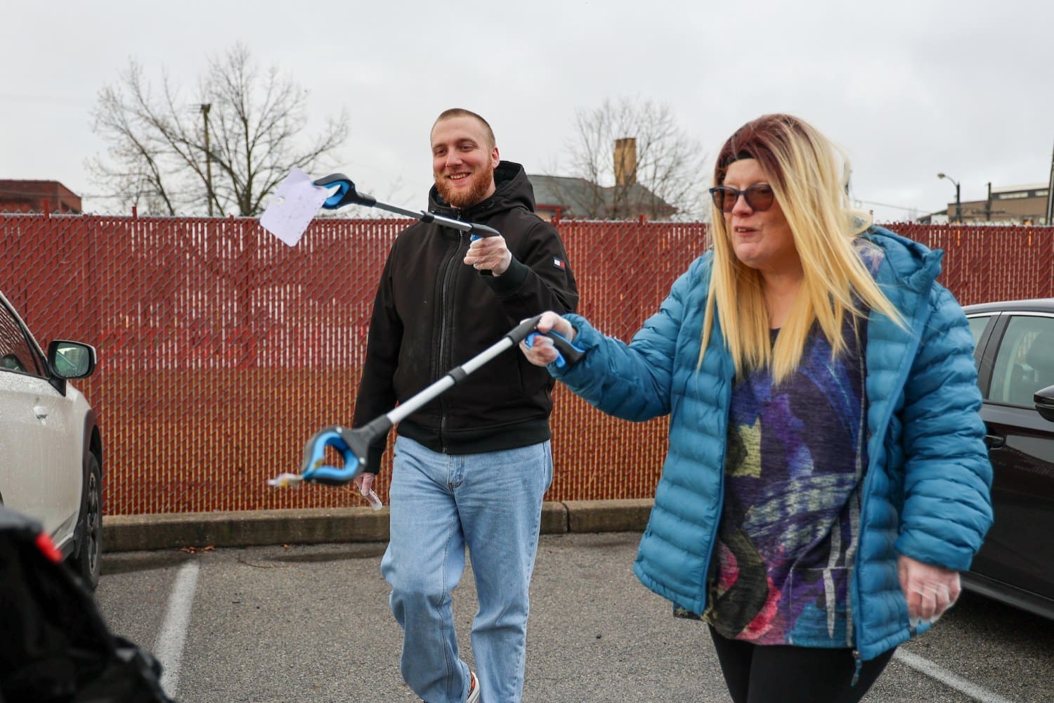 Two TLC individuals use trash pickers to clean up litter in a parking lot, wearing gloves and warm clothing on an overcast day.