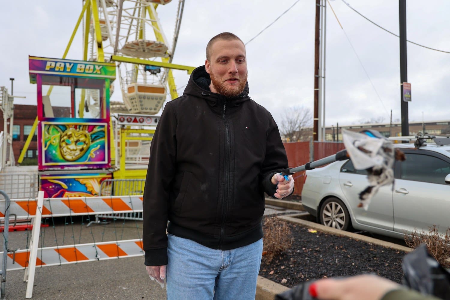 A TLC individual in a black hoodie uses a trash picker to collect litter near a carnival ride, placing it into a bag during a cleanup effort.