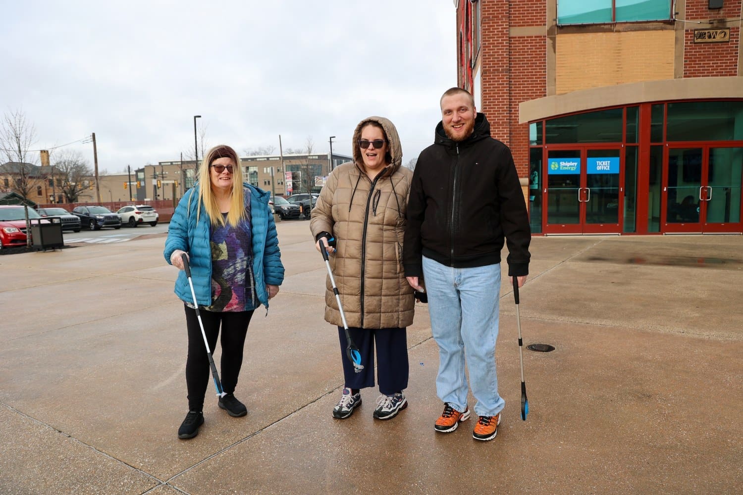 Three TLC individuals stand together outside a brick building, holding trash pickers and smiling during a community cleanup effort.
