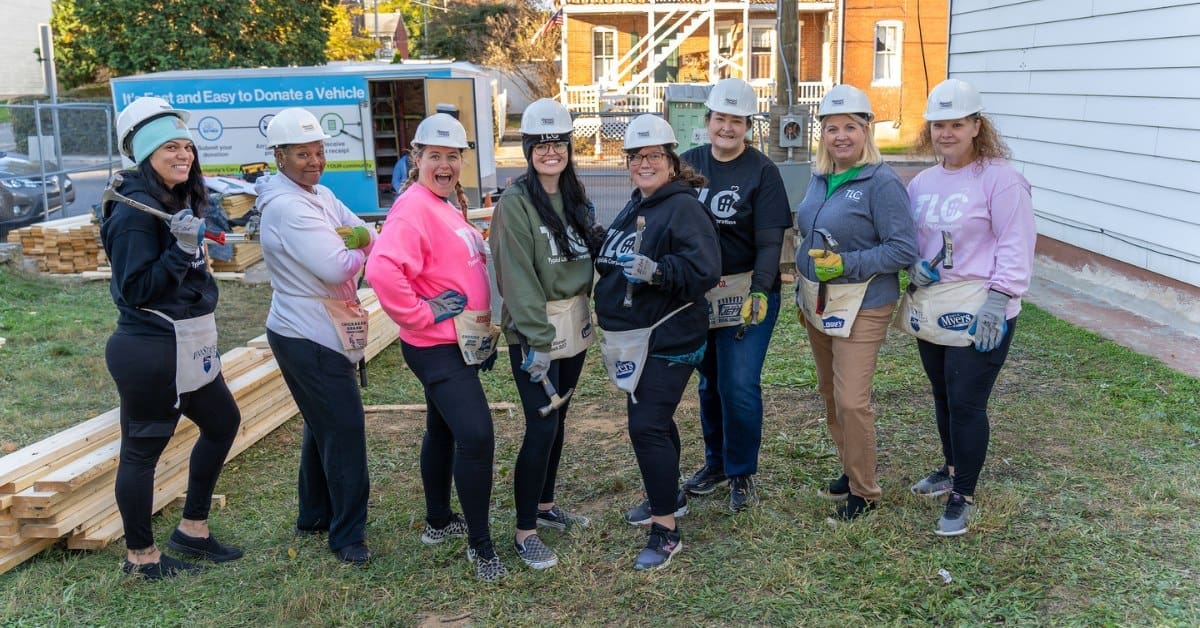 A group of TLC volunteers in hard hats and tool aprons pose together at a Habitat for Humanity construction site, smiling and holding tools.