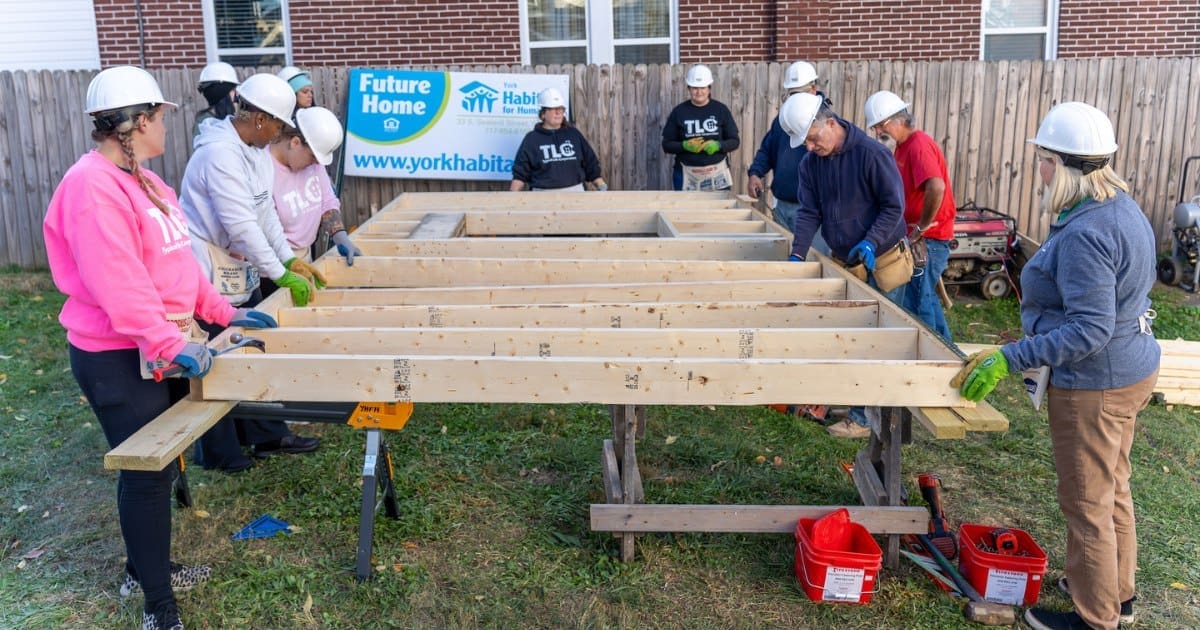 A group of TLC volunteers in hard hats and gloves work together to assemble a wooden frame at a Habitat for Humanity construction site.