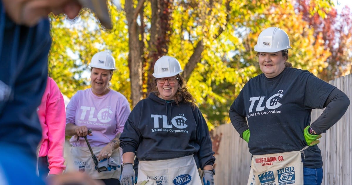 Three TLC volunteers in hard hats and tool aprons smile while participating in a Habitat for Humanity construction project.