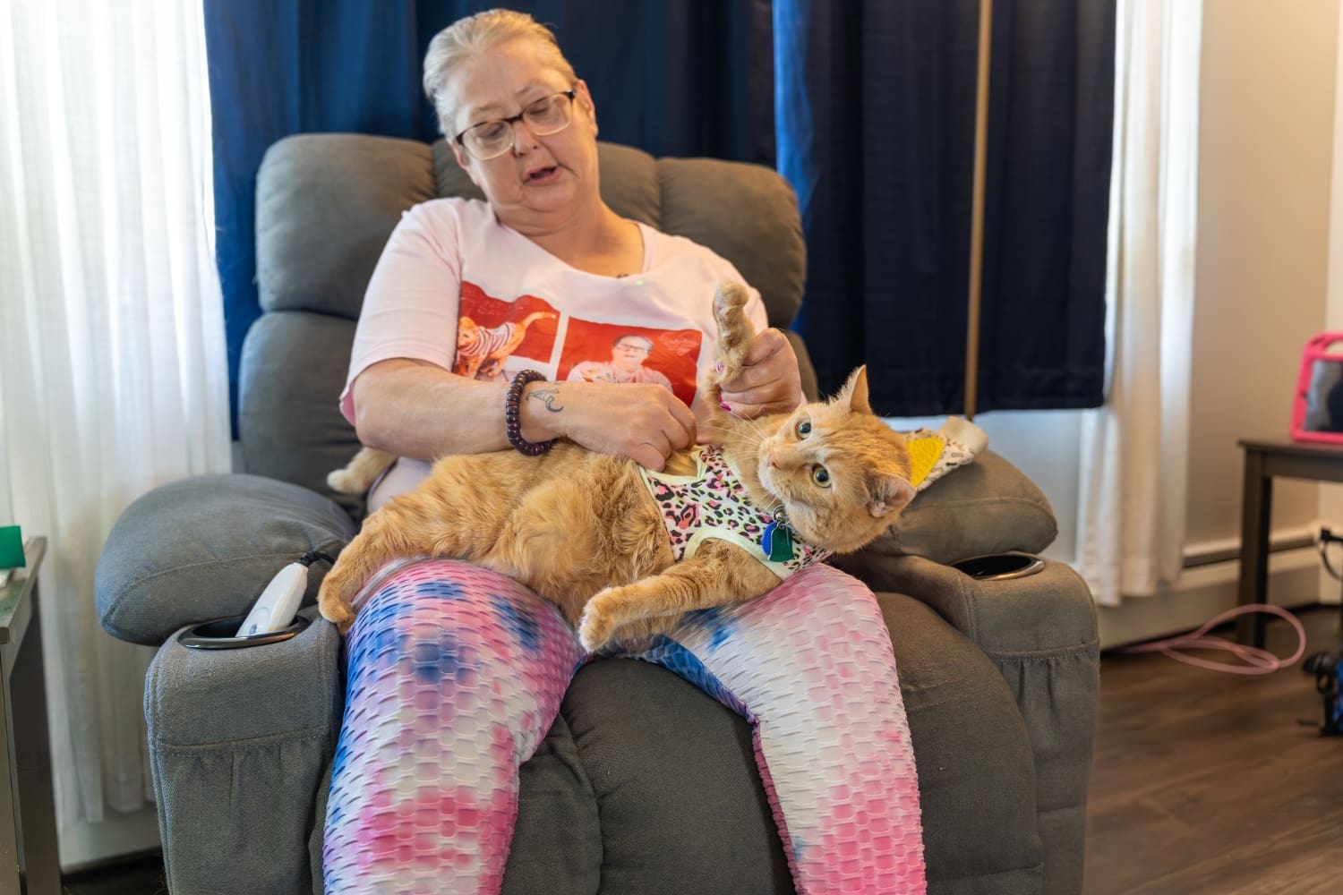 TLC resident Charro sits in a recliner with her orange cat, Buttercup, stretched across her lap wearing a colorful harness.